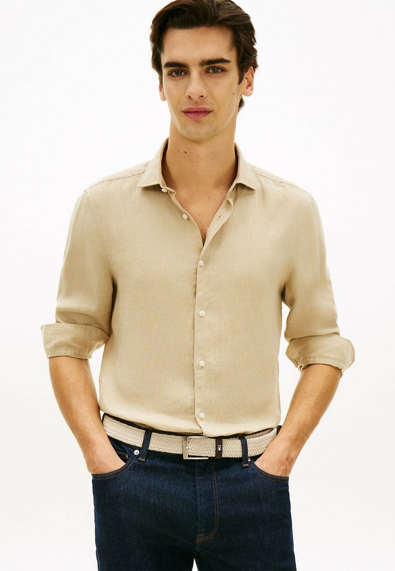 Jeune homme aux cheveux foncés portant une chemise beige à boutons avec manches retroussées, un jean sombre et une ceinture tissée beige, les mains dans les poches.