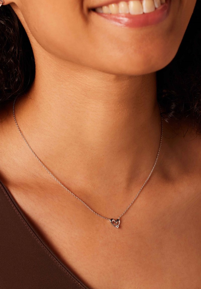 Collier en argent avec un pendentif en forme de cœur arborant une petite pierre précieuse, reposant sur une peau nue, avec un design de chaîne délicat.