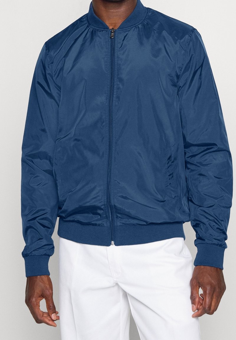 Homme portant une veste bomber bleu marine zippée avec col, poignets et ourlet côtelés, associée à un pantalon blanc.