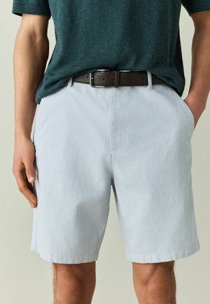 Mann trägt texturierte hellgraue Shorts mit Seitentaschen, einen dunkelbraunen Gürtel und ein eingestecktes dunkelgrünes Kurzarmhemd.