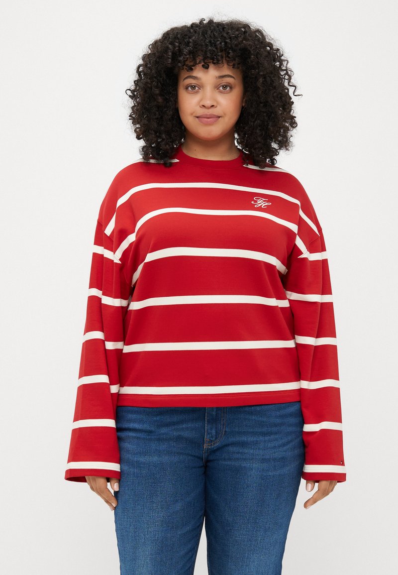 Maglietta rossa a maniche lunghe con strisce bianche orizzontali, caratterizzata da un collo a giro e logo ricamato sul petto, abbinata a jeans blu.