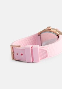 Correa de reloj de silicona rosa con superficie texturizada, que cuenta con un cierre y caja de metal color oro rosa, y una esfera de reloj transparente.