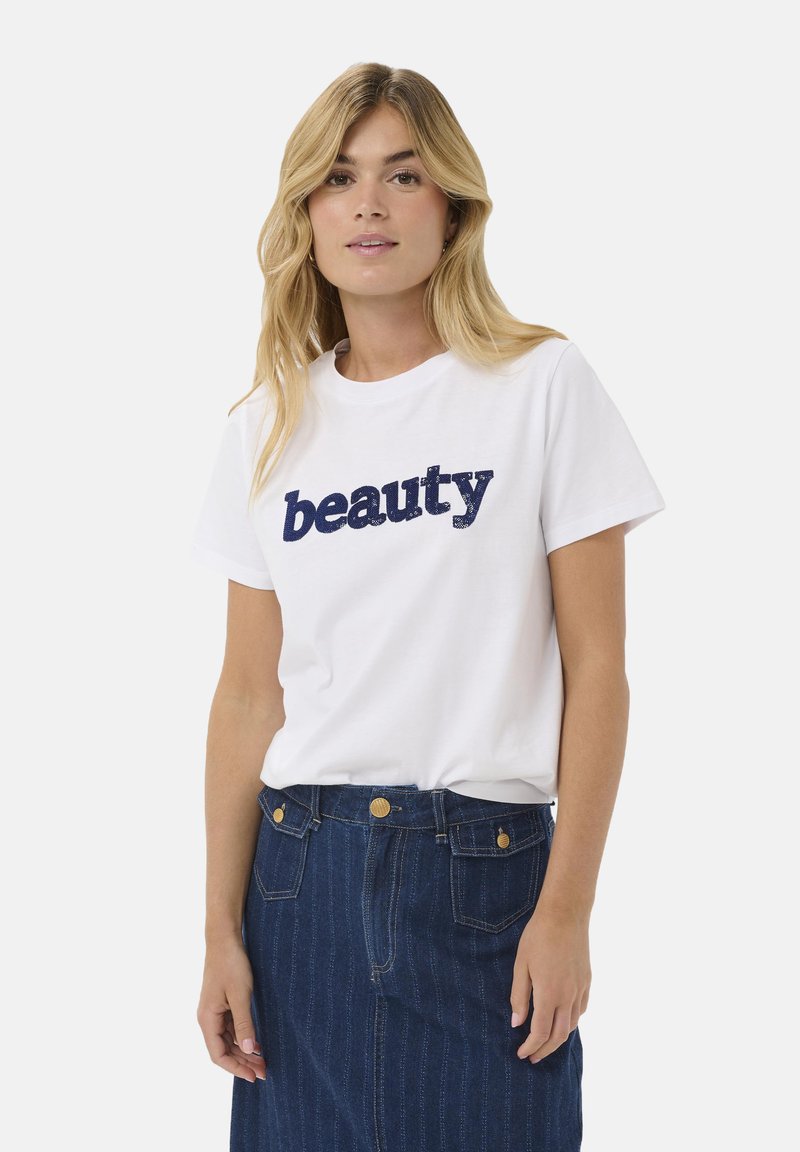 T-shirt en coton blanc avec un texte en paillettes bleues indiquant "beauté". Associé à une jupe en denim bleu foncé avec des coutures visibles et des poches.
