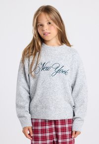 Pull gris à manches longues avec "New York" brodé en teal. Poignets et ourlet côtelés. Porté avec un pantalon écossais rouge. Tissu texturé doux.