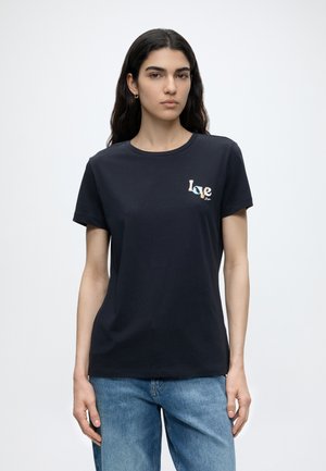 Mujer con cabello largo y oscuro lleva una camiseta azul marino con el texto "Love" en el pecho y jeans azul claro, de pie contra un fondo blanco liso.