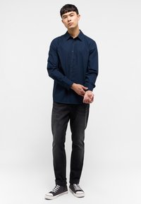 Camisa azul de botones con mangas largas, cuello clásico y un sutil logotipo. Combinada con jeans oscuros y zapatillas de suela blanca.