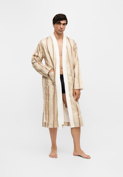 Homme non rasé aux cheveux foncés portant un peignoir beige jusqu'aux genoux avec des rayures verticales, debout pieds nus devant un fond blanc uni.