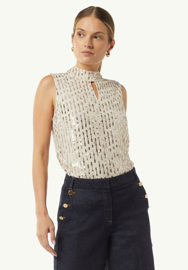 Paillettenoberteil, hoher Kragen, Schlüsselhemdetail, beige Farbe, vertikales Streifenmuster. Kombiniert mit dunkelblauen, hoch geschnittenen Jeans und goldenen Akzenten.