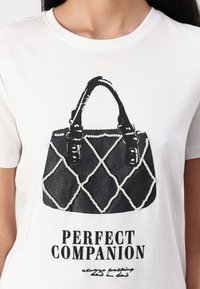 Hvit T-skjorte med grafikk av en svart håndveske med perledetaljer og quiltemønster, merket "PERFEKT FØLGE" under.