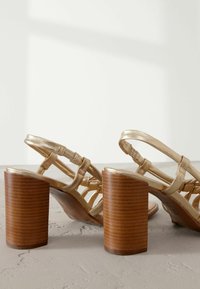 Guldremmede sandaler med blokhæl, der har en træagtig overflade og metalliske stropper, som fremhæver et elegant og minimalistisk design.