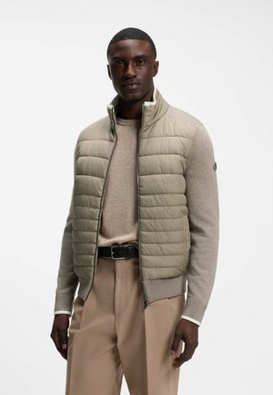 Mann trägt eine hellbeige gesteppte Weste über einem beigen Pullover mit beigen Hosen und schwarzem Gürtel, stehend vor weißem Hintergrund.