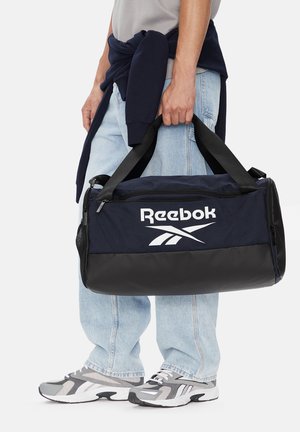 Osoba nosząca jasnoniebieskie dżinsy i szare trampki, trzymająca torbę sportową Reebok w kolorze granatowym i czarnym za uchwyty.