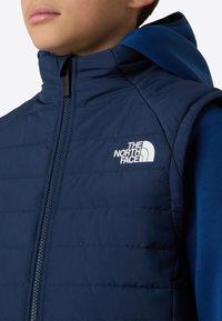 Vestă căptușită bleumarin cu guler ridicat și închidere cu fermoar, având un logo alb pe partea stângă a pieptului și o glugă interioară albastră contrastantă.