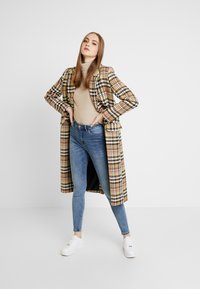 Νεαρή γυναίκα đứng με τα χέρια στη μέση, φορώντας ένα πολύχρωμο καμπαρντίνα με καρό σχέδιο, μπεζ ζιβάγκο, μπλε τζιν και λευκά αθλητικά παπούτσια.