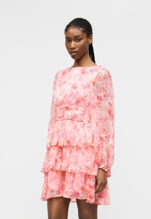 Kvinde med fletninger iført en langærmet, lyserød blomsterlaget kjole, stående mod en ensartet lys grå baggrund.