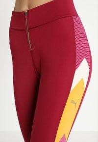 Leggings sportivi rossi con dettagli con zip, pannelli in rete rosa e un motivo geometrico con accenti gialli e bianchi.