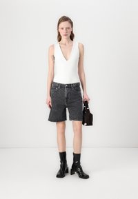 Bodysuit blanc sans manches avec décolleté en V plongeant, associé à un short en denim noir taille haute. Le modèle tient un petit sac à main marron, portant des bottes à talons noires.
