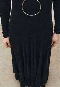 Schwarzes Kleid mit langen Ärmeln, das ein kreisförmiges silbernes Akzent am Taillenbereich und flared Falten hat, die von den Hüften nach unten verlaufen.