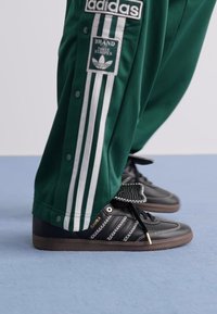 Grønne Adidas treningsbukser med hvite striper og logoer, iført sammen med svarte Adidas Samba-sneakers på et lyseblått gulv.