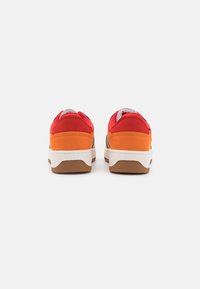 Zapatillas de ante rojas y naranjas con una entresuela blanca y suela de goma marrón, que presentan una lengua gris y acentos azules en el talón.