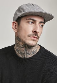 Grå ullcaps med flat brem, teksturert overflate, brukt på skrå. Svart strikkegenser med ribbestrikket mønster. Synlige nakke-tatoveringer.