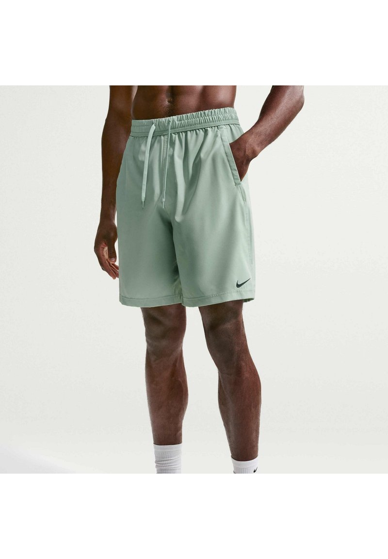Homme portant un short de sport Nike vert clair avec cordon de serrage, main dans la poche, et des chaussettes blanches Nike sur fond uni.
