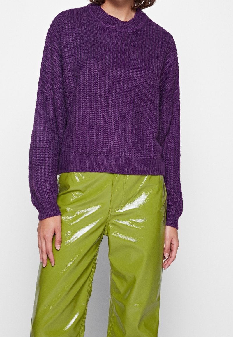 Femme portant un pull violet texturé à manches longues et un pantalon vert citron brillant sur un fond blanc uni.
