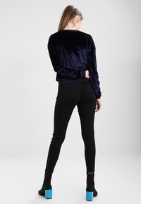 Maglietta lunga in velluto blu navy, jeans neri aderenti e stivaletti neri con tacco quadrato blu, visti da dietro.