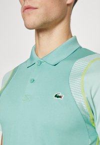 Polo shirt turquoise léger avec un col classique, deux boutons et des panneaux d'épaule texturés contrastants. Présente un petit logo Lacoste.