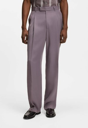 Hombre con pantalones morados de talle alto y sueltos con pliegues, mocasines negros y una camisa de rayas metida por dentro, de pie sobre fondo blanco.