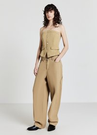 Frau mit lockigem, dunklem Haar trägt ein beiges trägerloses, geknöpftes Oberteil und weite Hosen, steht mit einer Hand in der Tasche und schwarzen Schuhen.