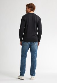 Zwarte langemouwen shirt met een ronde hals, gecombineerd met blauwe spijkerbroek en witte sportschoenen; zichtbare achterzakdetails.