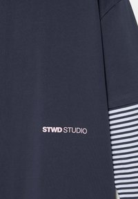 Cămașă bleumarin cu textul „STWD STUDIO” în roz pal, asortată cu un top cu mânecă lungă cu dungi bleumarin și albe dedesubt.