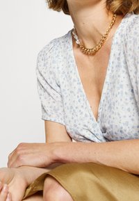 Blusa floral de color azul claro con mangas cortas, escote en V y un patrón texturizado, combinada con una cadena de oro y un collar de perlas.