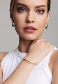 Złota bransoletka w formie bangle na nadgarstku, gładka faktura; modelka nosi kolczyki w kształcie kół. Biała bluzka z prążkami i neutralne tło. Zbliżenie na biżuterię.