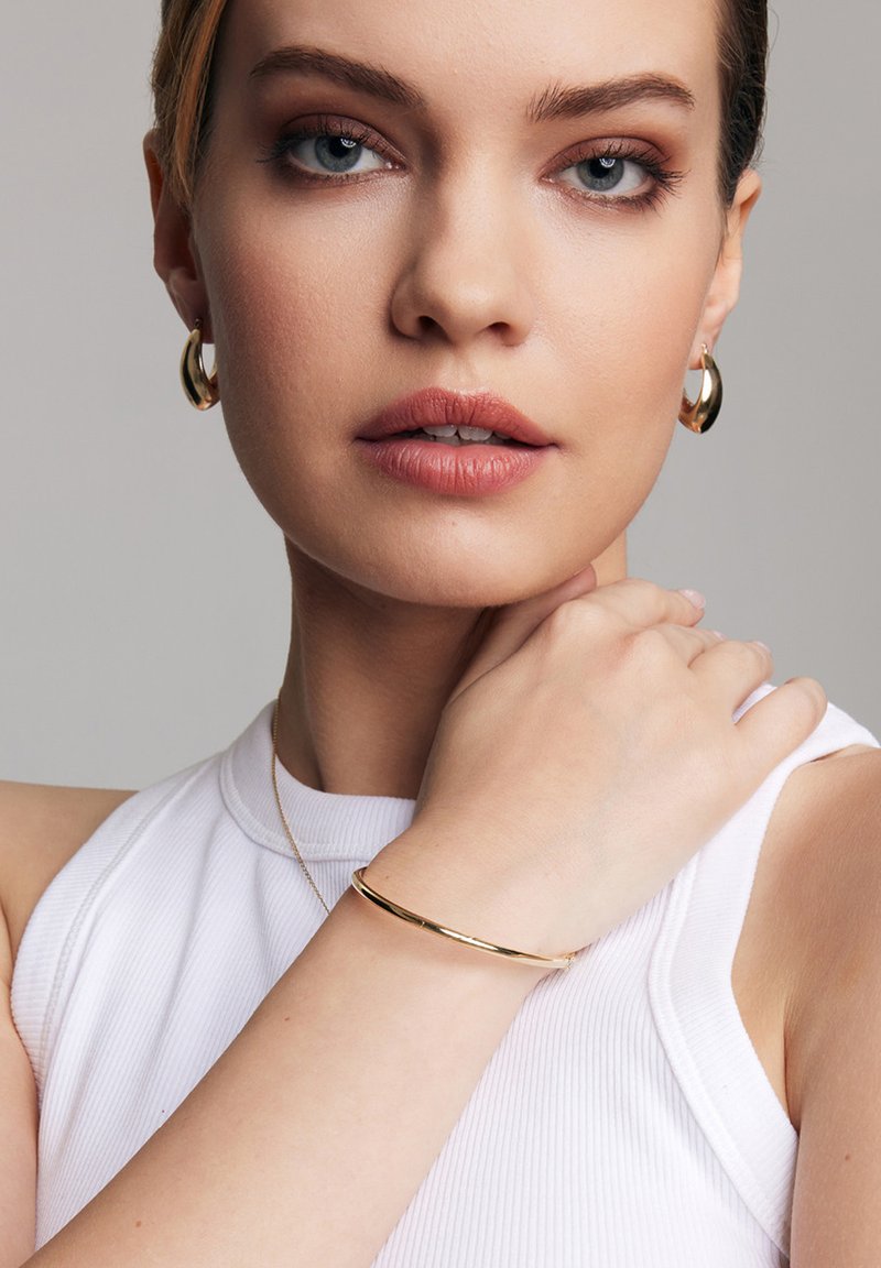 Złota bransoletka w formie bangle na nadgarstku, gładka faktura; modelka nosi kolczyki w kształcie kół. Biała bluzka z prążkami i neutralne tło. Zbliżenie na biżuterię.