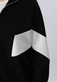 Veste noire avec une large bande horizontale blanche sur la partie supérieure de la manche et l'épaule, fermeture éclair au niveau du col.