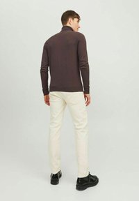 Maglione a collo alto marrone realizzato in un materiale a maglia aderente, abbinato a pantaloni color crema e scarpe nere. Modello visto da dietro.