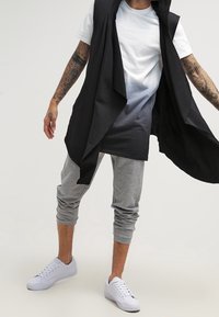 Man som bär en svart ärmlös öppen väst, en vit och svart ombre t-shirt, grå joggerbyxor, vita sneakers och synliga tatuerade armar.