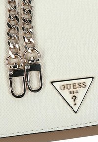 Weiße strukturierte Oberfläche einer Guess-Handtasche mit silbernen Kettenriemen und einer dreieckigen Guess USA-Logo-Plakette mit einem Fragezeichen.