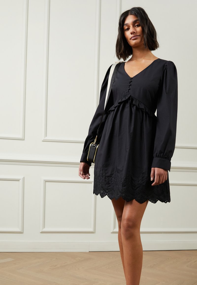 Vestido preto com mangas longas, frente abotoada, corpete com babados e barra de renda com extremidades recortadas. Usado com uma pequena bolsa preta.