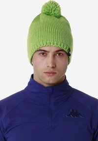 Berretto di lana verde con pompon sulla cima. Presenta una texture a coste e una piccola etichetta nera del brand sul lato. Indossato con una giacca azzurra con zip.