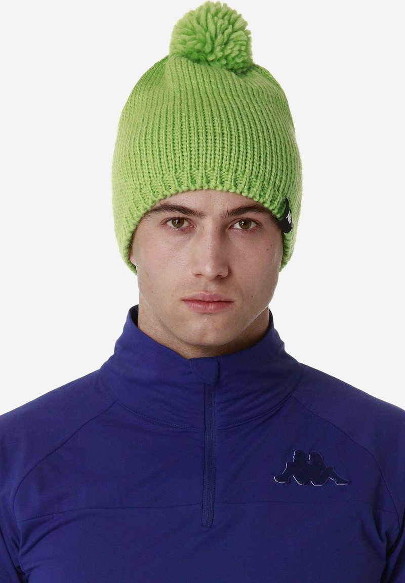 Berretto di lana verde con pompon sulla cima. Presenta una texture a coste e una piccola etichetta nera del brand sul lato. Indossato con una giacca azzurra con zip.