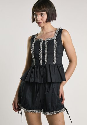 Vrouw draagt zwart mouwloos topje en korte broek met witte kanten rand, kijkt iets opzij tegen een effen achtergrond.