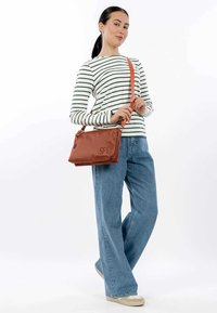 Brun textured håndtaske med justerbar orange skulderrem. Modellen har en stribet skjorte og vidde jeans i blå, og står foran en hvid baggrund.