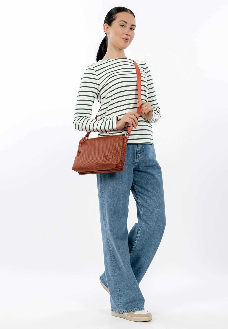 Brun textured håndtaske med justerbar orange skulderrem. Modellen har en stribet skjorte og vidde jeans i blå, og står foran en hvid baggrund.