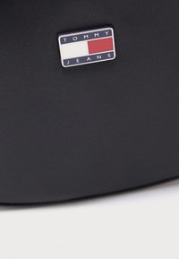 Fekete texturált kézitáska, melyen egy téglalap alakú fém logó található, rajta a "TOMMY JEANS" felirattal és piros, fehér, kék színű blokkokkal díszítve.