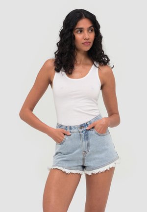 Vrouw met krullend zwart haar, gekleed in een wit tanktopje en lichtblauwe high-waisted denim shorts met rafelige randen, handen in de zakken.