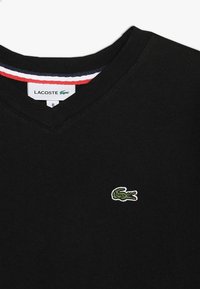 Czarny sweter w serek z bawełny. Posiada haftowane, zielone logo krokodyla na klatce piersiowej oraz w paski wykończenie na dekolcie w kolorach czerwonym, białym i niebieskim.