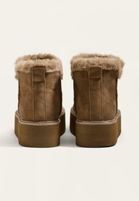 Brązowe zamszowe botki za kostkę z sztucznym futerkiem pod kołnierzem, grubą podeszwą i teksturowaną powierzchnią. Widok z tyłu, ukazujący detale projektu.
